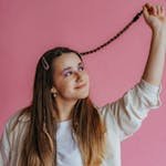 Smiling young woman holding braided hair against a pink backdrop in a studio setting.